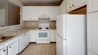 This single-floor residence offers a functional, well-organized layout across its living, sleeping, kitchen, and bathroom areas. The living room is a generously sized, carpeted space finished in neutral gray-toned walls, featuring a wide window that brings in natural light and provides views of the surrounding neighborhood. A wall-unit air conditioner and baseboard heating ensure year-round comfort, while a partial white countertop bar visually connects the living area to the kitchen.

The kitchen is designed with practicality in mind, featuring classic white cabinetry with ample upper and lower storage, granite-style countertops, and white appliances including a refrigerator, electric range, and dishwasher. Wood-look flooring runs throughout the kitchen, and recessed lighting adds brightness to the space. A pass-through opening connects the kitchen to the living area, enhancing the open feel.

The bedrooms are carpeted and finished in soft neutral tones, each equipped with baseboard heating and windows that admit natural light. The bathroom includes a tiled tub and shower combination, a window for ventilation, wood-look flooring, and a baseboard heater, offering a clean and efficient design throughout.