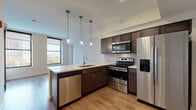 Located at 1 W Bowery St in Akron, Ohio, this single-floor unit offers a thoughtfully designed layout with modern finishes throughout. The kitchen serves as a central highlight, featuring dark espresso cabinetry, subway tile backsplash, stainless steel appliances including a French door refrigerator, electric range, built-in microwave, and dishwasher. A peninsula with a white countertop and undermount sink provides additional prep space and casual seating potential, while three pendant lights hang above, adding an elegant, contemporary touch. Light wood-tone flooring runs throughout the main living area, complementing the bright, neutral walls and creating a cohesive, open feel. Large windows allow natural light to filter in, offering views of the surrounding cityscape. The bedroom features plush wall-to-wall carpeting, a ceiling fan with integrated lighting, and a generously sized window that continues the theme of natural light. The bathroom is finished with large-format tile in the tub/shower enclosure fitted with sliding glass doors, a dark wood vanity topped with a white stone-style surface, a backlit mirror, and neutral floor tile, giving the space a clean, spa-like aesthetic.