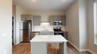 Located at 229 Roberts St N in Fargo, North Dakota, this single-floor unit offers a clean, modern aesthetic with thoughtfully designed living spaces. The open-concept layout connects the living area to a well-appointed kitchen, creating a functional and inviting flow throughout the unit. Warm-toned wood-look flooring runs throughout the main living areas, complementing the neutral wall palette.

The kitchen is a standout feature, showcasing grey shaker-style cabinetry with matte black hardware, crisp white quartz countertops, and a herringbone-patterned tile backsplash. A central island provides additional prep and seating space, while stainless steel appliances — including a refrigerator, electric range, built-in microwave, and dishwasher — complete the modern look. A double-basin sink with a contemporary faucet and a window offering natural light add to the kitchen's practicality and appeal.

The bathroom is finished with marble-look tile flooring in a bright white palette, a shaker-style vanity with a white countertop, black hardware accents, and a full tub-shower combination. Recessed lighting throughout the unit provides consistent, clean illumination. The overall design emphasizes a contemporary, move-in-ready feel with quality finishes and a cohesive neutral color scheme.