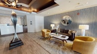 Located at 34 S Lexington Ave in White Plains, NY, this stylish single-floor lobby space offers a refined and welcoming environment defined by thoughtful design and high-end finishes. Wide-plank wood-look flooring extends throughout, creating a warm and cohesive foundation across the entire space.

The lounge area serves as a central focal point, featuring a rich brown leather sofa flanked by curvaceous golden yellow accent chairs and a sculptural marble-topped coffee table. A textured, steel-blue accent wall provides a dramatic backdrop, adorned with an ornate circular mosaic mirror that adds visual depth and artistry. Dual floor lamps with white drum shades offer balanced, ambient lighting throughout the seating arrangement.

Architectural interest is added through a striking coffered ceiling with warm copper-toned insets and recessed lighting, creating a layered and dramatic overhead element. Decorative wall panels with a honeycomb metallic pattern further enhance the lobby's contemporary character. The entry vestibule features modern glass and metal doors that invite natural light into the space.

Additional areas include a reception counter, a small refreshment station, private office or service doors, and elevator access — all finished with consistent trim details and neutral tones. Colorful artwork displayed on the walls contributes to the space's polished, gallery-like atmosphere.