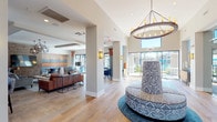 This thoughtfully designed single-story community clubhouse blends contemporary and transitional styles across a spacious, open-concept floor plan. The entry area is anchored by a striking round statement chandelier with candle-style lighting and a decorative upholstered ottoman, all set atop light-toned hardwood flooring that extends throughout the main gathering areas.

The central lounge features comfortable seating arrangements, including leather sofas and velvet accent chairs, positioned beneath bold geometric pendant fixtures. Adjacent to the lounge, a demonstration kitchen showcases light wood cabinetry, dark countertops, a large island with bar seating, and pendant lighting with an artisan glass finish.

A dedicated dining and social zone features exposed brick accent walls, wood-paneled TV display, and high-top bar seating, adding a warm, urban character. Nearby, a casual dining nook with upholstered banquette seating and round tables is framed by floor-to-ceiling sliding glass doors dressed in two-tone curtains. These doors open directly onto a covered outdoor patio with lounge seating, umbrellas, and views of a resort-style pool, seamlessly connecting the interior to the exterior entertaining spaces.