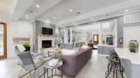 This single-story community clubhouse is designed with a sophisticated yet welcoming aesthetic throughout. The grand entrance lobby sets the tone with soaring tray ceilings, a statement globe chandelier, herringbone-patterned light stone flooring, and decorative columns that define the space without obstructing sightlines. A cowhide rug and live-edge accent table add organic warmth to the airy entryway.

The open lounge area flows naturally from the lobby and serves as the social heart of the space. A stacked stone accent wall frames a classic fireplace with a white mantel and mounted flat-screen television, while plush seating invites relaxation. Recessed lighting and crown molding enhance the refined atmosphere.

Adjacent to the lounge is a fully functional kitchen featuring rich dark wood cabinetry, quartz countertops, a mosaic tile backsplash, stainless steel appliances, and a central island with bar seating — ideal for community gatherings and events.

The business center offers a quiet, carpeted workspace with a built-in desk, overhead cabinetry, and generous natural light from large windows with wood blinds. A dedicated fitness center, enclosed by floor-to-ceiling glass walls, is outfitted with cardio machines, strength training equipment, free weights, medicine balls, and exercise balls beneath a tray ceiling with a ceiling fan.