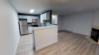 Located at 12825 60th Ln S, Unit B4, in Seattle, WA 98178, this single-floor unit features a thoughtfully designed open-concept layout that seamlessly connects the kitchen and living area. The kitchen is well-appointed with white shaker-style cabinetry, stainless steel appliances including a refrigerator and electric range with overhead microwave, and a peninsula that provides additional counter space while creating a natural division between the kitchen and the main living area. The neutral blue-gray accent walls complement the crisp white cabinetry and trim, lending the space a clean, contemporary feel. Light wood-look flooring runs throughout, enhancing the sense of openness and brightness. A recessed ceiling light fixture in the kitchen ensures ample task lighting, while a baseboard heater along the living room wall provides efficient heating. The textured ceiling adds a subtle layer of character to the interior. Overall, the space presents a modern, move-in-ready environment with a cohesive color palette and practical finishes suited for comfortable everyday living.
