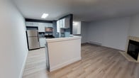Located at 12825 60th Ln S, Unit B4, in Seattle, WA 98178, this single-floor unit features a thoughtfully designed open-concept layout that seamlessly connects the kitchen and living area. The kitchen is well-appointed with white shaker-style cabinetry, stainless steel appliances including a refrigerator and electric range with overhead microwave, and a peninsula that provides additional counter space while creating a natural division between the kitchen and the main living area. The neutral blue-gray accent walls complement the crisp white cabinetry and trim, lending the space a clean, contemporary feel. Light wood-look flooring runs throughout, enhancing the sense of openness and brightness. A recessed ceiling light fixture in the kitchen ensures ample task lighting, while a baseboard heater along the living room wall provides efficient heating. The textured ceiling adds a subtle layer of character to the interior. Overall, the space presents a modern, move-in-ready environment with a cohesive color palette and practical finishes suited for comfortable everyday living.