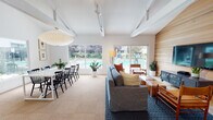 Located at 3770 Flora Vista Ave in Santa Clara, CA, this single-story community clubhouse offers a thoughtfully designed gathering space with a welcoming, mid-century modern aesthetic. The open-plan main area seamlessly integrates a lounge and dining zone, bathed in abundant natural light streaming through large picture windows and sliding glass doors that frame direct views of the outdoor pool and surrounding greenery. Exposed ceiling beams and track lighting add architectural character overhead, while a striking horizontal wood-clad accent wall anchors the lounge area, complete with a wall-mounted television and comfortable seating arrangements. The dining section accommodates a generous long table with ample seating, illuminated by a classic pendant light fixture. A galley-style kitchen features white shaker cabinetry with matte black hardware, sleek grey countertops, subway tile backsplash, and dark slate floor tiles — providing a clean, functional space for event catering or community gatherings. A private meeting room offers an intimate setting with deep teal walls, a round wood table, and curated artwork, ideal for smaller group discussions. The entry foyer is bright and open, featuring vaulted ceilings, decorative artwork, and dual glass doors that connect seamlessly to the outdoor landscape.
