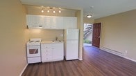 This single-floor space combines a functional kitchen and open living area in a clean, practical layout. The kitchen is outfitted with white shaker-style cabinetry extending to the ceiling, providing ample storage. White appliances — including a freestanding electric range and a full-size refrigerator — complement the bright cabinetry, while a single-basin sink with a brushed metal faucet sits beneath the counter. A track lighting fixture with multiple heads illuminates the kitchen workspace effectively. The flooring throughout is a rich, dark-toned hardwood that adds warmth and visual contrast to the light-colored walls and cabinetry. The open layout transitions naturally from the kitchen into the living area, which benefits from natural light streaming through a glazed panel beside the entry door. The solid, dark red entry door makes a bold focal point and opens to what appears to be an outdoor common area, with a staircase visible just beyond, suggesting a multi-unit building. The warm beige wall tones and flush ceiling light in the living area contribute to an inviting, neutral ambiance throughout the space.