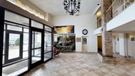 This single-floor lobby makes an immediate impression with its generous ceiling height, creating an open and airy atmosphere upon entry. Large glass doors at the entrance allow natural light to flow freely into the space, brightening the neutral-toned ceramic tile flooring that extends throughout. A decorative wrought-iron chandelier serves as a focal point overhead, adding a touch of elegance to the welcoming environment.

The focal wall features a vivid, large-format photographic mural depicting a natural waterfall and rocky landscape, paired with a dramatic sunset canvas artwork displayed above it, lending the space a nature-inspired aesthetic. A rustic stone pillar accent on one side of the lobby complements these natural motifs and adds textural contrast to the otherwise smooth white walls.

A traditional wall clock is mounted near a secondary doorway, adding a functional yet classic detail to the space. A single armchair is positioned near the entrance, providing a seating option for visitors. An upper-level balcony or mezzanine is visible in the background, hinting at additional space above. Overall, the lobby balances practicality with decorative charm, creating a comfortable and visually engaging reception area.