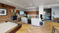 This single-floor communal space is thoughtfully designed to balance leisure and socializing across several distinct yet open zones. The standout flooring throughout is a striking chevron-patterned hardwood in a warm natural tone, lending a sense of movement and visual continuity across the entire space.

At the heart of the recreation area sits a substantial pool table, positioned beneath recessed ceiling lighting that highlights the room's generous proportions. A rich, leather-paneled accent wall serves as a sophisticated backdrop, adorned with framed black-and-white artwork. Large floor-to-ceiling sliding glass doors open directly to an outdoor terrace, flooding the interior with natural light and providing a seamless indoor-outdoor connection.

Adjacent to the game area is a comfortable lounge zone featuring upholstered seating, a wall-mounted flat-screen display, and carefully curated décor including potted plants and decorative accessories. A compact wet bar with stainless steel appliances offers added convenience for entertaining.

A more intimate dining nook is tucked alongside the terrace access, furnished with a dark banquette sofa accented by colorful throw pillows, a round café table, and wicker chairs — its patterned wall treatment adding a distinctive decorative detail. Recessed lighting throughout ensures a warm, inviting ambiance across all areas.