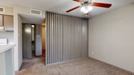 This single-floor residential unit offers a practical and straightforward layout designed for comfortable everyday living. The kitchen is positioned near the entry and features a peninsula counter that separates it from the living area, providing both additional workspace and a casual bar-style seating option. Cabinetry in a neutral taupe finish lines the walls, complemented by white appliances including a refrigerator, electric range, and dishwasher. Wood-look flooring runs through the kitchen, transitioning to carpet in the living spaces. A distinctive feature throughout the living and bedroom areas is a full-height slatted wall panel with a vertical ribbed texture, lending a retro architectural character to the interior. Carpeted floors in a warm beige tone carry through these rooms, adding softness underfoot. Ceiling fans with integrated light fixtures are present in the main rooms, providing both ventilation and ambient lighting. The textured popcorn ceilings are consistent throughout. The bathroom includes a full-size soaking tub and shower combination with clean white tile surrounds and chrome fixtures. Overall, the unit presents a neutral, functional palette with a few distinctive design details that give it a recognizable character.