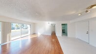 This single-floor unit offers a clean, well-lit interior defined by warm hardwood-style flooring that flows throughout the main living areas and bedrooms, complemented by light tile in the kitchen and bathroom. The expansive open living and dining area is a standout feature, bathed in natural light streaming through large sliding glass doors and multiple windows that frame views of the surrounding neighborhood. A textured popcorn ceiling adds character throughout the main living spaces. The kitchen is thoughtfully appointed with white shaker-style cabinetry, ample counter space, a stainless steel dishwasher, and a smooth-top range, all set over clean light tile flooring. The layout, as revealed by the dollhouse view, includes multiple rooms connected by a central hallway, providing a functional and organized flow. The bedrooms are generously sized, each featuring hardwood-style flooring and bifold closet doors offering built-in storage. The full bathroom is finished in an all-white palette, complete with a tiled shower-tub combination, a vanity with sink, and tile flooring. Throughout the unit, crisp white walls and consistent finishes create a bright, neutral, and move-in ready environment.