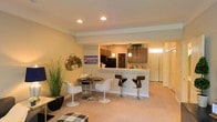 This two-floor residence offers a thoughtfully designed open-concept layout that effortlessly connects the living, dining, and kitchen areas into one cohesive and functional space. The main living area is finished with warm, neutral-toned carpeting that adds a sense of comfort and continuity throughout the space. Recessed lighting in the ceiling provides even, ambient illumination, complemented by decorative pendant lights that hang above the kitchen bar area, adding a stylish focal point. The kitchen features dark wood cabinetry, black appliances, and a raised breakfast bar equipped with modern swivel bar stools, making it ideal for casual dining and entertaining. A compact dining nook with a round table and contemporary tulip-style chairs sits adjacent to the kitchen, offering an intimate space for meals. Decorative wall art and lush indoor greenery contribute to the warm, inviting ambiance of the main level. Crown molding along the ceiling adds an elegant architectural detail that elevates the overall aesthetic. A secondary entrance door with a sidelite window brings in natural light near the kitchen. With two floors of living space, this home provides ample room for both relaxation and everyday living.
