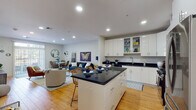 Located at 1779 Central St in Stoughton, MA, this single-floor residence offers a thoughtfully designed open-concept layout with warm hardwood flooring throughout. The kitchen is a standout feature, boasting high-gloss white cabinetry, dark granite countertops, stainless steel appliances including a gas range, built-in microwave, and refrigerator, and a central island with bar seating — ideal for both cooking and casual dining. The kitchen flows seamlessly into a bright living area, where recessed lighting and neutral walls create a clean, welcoming atmosphere. A dedicated dining nook features a round white table and rattan chairs, accented by decorative wall-mounted planters. The living room offers generous space for multiple seating arrangements and connects to a secondary lounge area with wall-mounted television. The bedroom is a serene retreat, highlighted by a bold teal accent wall, natural light from a window with outdoor views, and a walk-in closet. The bathroom is finished with large-format white tile flooring, a full vanity with white cabinetry, a combined tub and shower with glass sliding doors, and built-in shelving for added storage. Recessed lighting throughout enhances the clean, modern aesthetic of this well-appointed home.