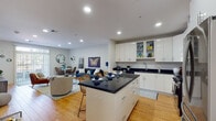 Located at 1779 Central St in Stoughton, MA, this single-floor residence offers a thoughtfully designed open-concept layout with warm hardwood flooring throughout. The kitchen is a standout feature, boasting high-gloss white cabinetry, dark granite countertops, stainless steel appliances including a gas range, built-in microwave, and refrigerator, and a central island with bar seating — ideal for both cooking and casual dining. The kitchen flows seamlessly into a bright living area, where recessed lighting and neutral walls create a clean, welcoming atmosphere. A dedicated dining nook features a round white table and rattan chairs, accented by decorative wall-mounted planters. The living room offers generous space for multiple seating arrangements and connects to a secondary lounge area with wall-mounted television. The bedroom is a serene retreat, highlighted by a bold teal accent wall, natural light from a window with outdoor views, and a walk-in closet. The bathroom is finished with large-format white tile flooring, a full vanity with white cabinetry, a combined tub and shower with glass sliding doors, and built-in shelving for added storage. Recessed lighting throughout enhances the clean, modern aesthetic of this well-appointed home.