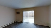 This single-floor space offers a straightforward and functional layout designed for comfortable living. The main room is finished with wall-to-wall carpeting in a neutral grey tone, providing a soft and practical underfoot surface. A prominent sliding glass door serves as the room's focal point, fitted with vertical blinds that allow for adjustable light control and privacy. The door opens to a view of a natural outdoor setting, with trees and open lawn visible beyond — a pleasant, calming backdrop that brings a sense of the outdoors inside. Natural light filters through the glass, brightening the otherwise simply appointed interior. The walls are finished in a warm beige tone, complementing the neutral color palette throughout. Climate control is provided by a wall-mounted air conditioning unit, while a baseboard heating system runs along the base of the exterior wall, ensuring year-round temperature management. The ceiling features a textured finish and sits at a standard height, contributing to the room's modest but functional character. Overall, this space presents a clean, no-frills environment with practical amenities and a pleasant connection to the surrounding natural landscape.