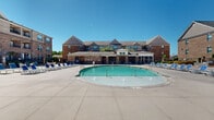 Located at Walkertown Landing in Walkertown, North Carolina, this two-story community showcases a well-appointed outdoor amenity area centered around a generously sized resort-style swimming pool. The pool features a smooth curvilinear design with built-in fountain jets and clearly marked depth gradations, surrounded by an expansive concrete deck lined with blue lounge chairs ideal for relaxing under the open sky.

The surrounding brick and stone architecture of the multi-story building provides a classic, cohesive backdrop to the outdoor space. A dedicated covered pavilion serves as the focal point of the poolside area, constructed with stacked stone columns and a coffered ceiling fitted with ceiling fans for warm-weather comfort. The pavilion includes a built-in bar counter with bar-height seating, offering a convenient gathering spot for residents.

Adjacent to the pavilion, an open patio area is furnished with cushioned outdoor lounge chairs and accent tables, providing a comfortable transition between the indoor clubhouse and the pool deck. Large glass storefront windows along the clubhouse facade allow natural light to flow freely and maintain visual connectivity between the interior and exterior spaces. Pavers and concrete surfaces throughout the deck area add textural variety and durability to the overall design.