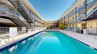 This outdoor pool area at 4701 E Mississippi Ave in Glendale, Colorado, serves as a central communal amenity within a multi-story residential complex. The space is defined by a generously sized rectangular swimming pool with clear, blue water, positioned at the heart of an open-air courtyard. Surrounding the pool is a spacious concrete sun deck furnished with lounge chairs and side tables, providing ample room for sunbathing and relaxation under Colorado's abundant sunshine.

A standout feature of the space is the covered outdoor lounge area, sheltered by a rustic wood-beam pergola with a corrugated metal ceiling and strung with overhead lighting. This shaded retreat is furnished with wicker high-top tables and bar chairs, offering a comfortable spot to gather while overlooking the pool. Vibrant flower planters with colorful blooms add a lively, welcoming touch to the poolside environment.

The courtyard is enclosed on multiple sides by the multi-level building, whose exterior features light brick facades, dark metal railings, and open-air balconies accessed by exterior stairways. Decorative rock landscaping and ornamental planters accent quieter corners of the deck. The open-sky setting ensures the area is bathed in natural light throughout the day, creating a bright and inviting outdoor retreat.