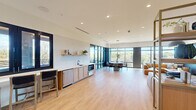 This contemporary amenity lounge is a thoughtfully designed, single-level space that seamlessly blends comfort and modern aesthetics. Light wood-look flooring runs throughout the open interior, connecting distinct functional zones while maintaining an airy, cohesive feel. The kitchenette area is anchored by warm wood cabinetry, a white subway tile backsplash with open floating shelves, a built-in sink, and under-counter appliances — ideal for entertaining or casual gatherings. A built-in bar-height counter with seating offers a dedicated workspace or casual dining spot alongside stylish open shelving displays. The lounge area features generous leather sectional sofas arranged beneath oversized dome pendant lights, with wall-mounted flat-screen televisions and curated artwork set against a richly textured blue-grey accent wall. Floor-to-ceiling windows flood the interior with natural light and frame views of a waterway and lush treetops beyond. Recessed ceiling lighting throughout ensures a well-lit environment at any hour. Accessible from the interior, a covered outdoor terrace extends the living space further, finished with large-format tile flooring, a warm wood-plank ceiling, an outdoor TV, and multiple seating arrangements — making it an inviting retreat to enjoy open-air views of the surrounding landscape.
