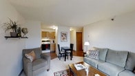 This single-floor unit offers an open-concept layout that seamlessly integrates the living room, dining area, and kitchen into one connected space. The living room is comfortably sized, featuring hardwood flooring partially covered by area rugs, and is furnished with a grey sofa set alongside a wooden coffee table, creating a relaxed and functional atmosphere. A textured ceiling adds subtle character throughout the space.

The compact dining area sits between the living room and kitchen, accommodating a dark-finished table with matching chairs — ideal for casual meals. Adjacent to it, the kitchen is efficiently designed with warm wood-toned cabinetry, a white gas range, and sufficient counter space for everyday cooking needs.

A hallway visible from the main living area leads to additional rooms, suggesting a practical floor plan that keeps private spaces separated from the common areas. The warm wood door at the hallway's end adds a classic touch to the interior. Overall, the space offers a straightforward, functional layout with warm tones and an open feel suitable for everyday living.