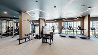 Located in Orlando, FL, this contemporary fitness center spans a single floor and is divided into two well-equipped gym areas, offering a comprehensive workout environment. The main gym area features striking hexagonal LED pendant lighting suspended from dark-painted ceilings, creating a sleek, modern atmosphere. Large floor-to-ceiling windows flood the space with natural light and provide views of the outdoor surroundings, including palm trees and a pool area. This section houses a multi-station cable machine, ellipticals, treadmills with personal screens, and a rowing machine, all set on neutral-toned carpet flooring. Full-length mirrors amplify the sense of space throughout. A dedicated studio room, enclosed by black-framed glass sliding doors, offers hardwood flooring, a wall-mounted flat-screen TV, dumbbell rack, and space for functional training. Decorative hexagonal wood wall panels and warm wood cabinetry with a built-in mini-refrigerator add refined touches near the studio entrance. A separate spin studio features multiple cycling bikes with digital displays, floor-to-ceiling mirrors, and large windows with outdoor views. The overall design balances high-end aesthetics with practical functionality, making it a well-appointed fitness facility.