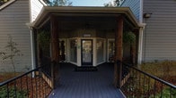 Located at 2000 Bay Area Blvd in Houston, Texas, this single-story leasing office serves as the welcoming hub for The Cove Apartments community. The exterior immediately sets a warm tone, featuring a covered front porch supported by rustic wood columns, a wood-plank deck, and large bay windows that frame the entrance and allow natural light to pour in.

Upon entry, visitors are greeted by a bright, open lobby area finished with warm hardwood-style flooring and light gray walls accented by a bold teal feature wall. A reception counter with a "Welcome" sign faces a comfortable seating area, creating an inviting atmosphere for prospective and current residents alike. Multiple windows with blinds line the space, ensuring the interior remains well-lit throughout the day.

Beyond the lobby, a dedicated staff office area is equipped with a work desk, storage cabinetry, and a secondary windowed workspace visible through an interior glass partition. The open-plan layout connects the reception zone to a broader community gathering area at the rear, which features exposed beam ceiling details, additional seating with dining-style furniture, and expansive windows that overlook the surrounding greenery.

Throughout the office, hardwood-style flooring, textured ceilings, and a carefully coordinated color palette of soft grays, teal, and warm wood tones contribute to a cohesive, professional yet approachable aesthetic.