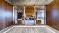 Located at 800 Main St in Hartford, CT, this elegantly designed high-rise space spans an expansive single floor, offering a seamlessly connected series of lounge, dining, and support areas. The entry foyer makes an immediate impression with polished marble flooring, rich wood-paneled walls, a textured grid ceiling, and a striking central gold sculptural fountain — setting a refined, gallery-like tone. Framed artwork lines the corridor walls, reinforcing the space's curated aesthetic. Through floor-to-ceiling glass doors, a sophisticated lounge area unfolds, anchored by a warm gold ceiling, deep blue velvet sofas, and a large marble accent wall that serves as a dramatic focal point. Adjacent to this, a more casual seating zone features bold red lacquered ceilings, plush rounded leather furnishings, and vivid patterned carpeting. Floor-to-ceiling windows throughout bathe the interiors in natural light and frame sweeping panoramic views of the Hartford skyline and beyond, with access to an outdoor terrace. A dining and gathering area offers sleek black tables paired with modern chairs beneath the same striking red ceiling. The support kitchen is outfitted with dark granite countertops, white cabinetry with brushed gold hardware, and tile flooring — functional yet refined. Together, these areas compose a versatile, design-forward space ideal for upscale events and gatherings.