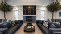 Located at 484 Greenmont Dr in Lewis Center, Ohio, this single-floor community facility combines contemporary design with inviting, functional spaces. The centerpiece of the space is a generously proportioned lounge area anchored by a striking floor-to-ceiling fireplace clad in dark stacked stone, accented with a bold black-and-gold geometric tile surround. Sleek black leather sofas arranged symmetrically create an elegant and comfortable seating arrangement, complemented by marble-top round coffee tables and herringbone-patterned hardwood flooring that runs throughout. Large windows on multiple walls flood the lounge with abundant natural light while offering views of the surrounding outdoor spaces. Crown molding and recessed lighting add refined architectural detail. A dedicated beverage station features crisp white shaker cabinetry, dramatic black granite countertops, a Keurig coffee maker, and a stainless steel beverage refrigerator — all topped by a wall-mounted flat-screen TV. The entry area presents a polished black reception desk framed by lush potted trees and vibrant floral arrangements, setting a sophisticated first impression. A corridor connects to an indoor pool area, rounding out this well-appointed community hub. Curated artwork and decorative greenery are displayed throughout, enhancing the upscale, welcoming ambiance of the entire space.