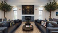 Located at 484 Greenmont Dr in Lewis Center, Ohio, this single-floor community facility combines contemporary design with inviting, functional spaces. The centerpiece of the space is a generously proportioned lounge area anchored by a striking floor-to-ceiling fireplace clad in dark stacked stone, accented with a bold black-and-gold geometric tile surround. Sleek black leather sofas arranged symmetrically create an elegant and comfortable seating arrangement, complemented by marble-top round coffee tables and herringbone-patterned hardwood flooring that runs throughout. Large windows on multiple walls flood the lounge with abundant natural light while offering views of the surrounding outdoor spaces. Crown molding and recessed lighting add refined architectural detail. A dedicated beverage station features crisp white shaker cabinetry, dramatic black granite countertops, a Keurig coffee maker, and a stainless steel beverage refrigerator — all topped by a wall-mounted flat-screen TV. The entry area presents a polished black reception desk framed by lush potted trees and vibrant floral arrangements, setting a sophisticated first impression. A corridor connects to an indoor pool area, rounding out this well-appointed community hub. Curated artwork and decorative greenery are displayed throughout, enhancing the upscale, welcoming ambiance of the entire space.