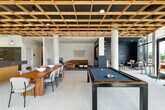 This thoughtfully designed amenity space offers a dynamic blend of social and relaxation areas across a single open floor. The architectural centerpiece is a striking wood-lattice ceiling grid that spans the main gathering area, adding warmth and visual texture while complementing the large-format tile flooring below. Floor-to-ceiling glass walls and doors flood the interior with natural light and frame sweeping views of the surrounding waterfront and outdoor terrace, creating a seamless indoor-outdoor connection.

The main space features a large kitchen island with a dark granite countertop and warm wood cabinetry, accompanied by pendant globe lighting. A full suite of appliances and colorful tile backsplashes — in both deep green and rich burgundy — add bold design accents. Nearby, a live-edge communal dining table provides ample gathering space, while a billiards table positioned beneath the lattice ceiling invites friendly competition.

A dedicated lounge area offers a more intimate setting, featuring a tufted caramel leather sectional, curated seating, and a built-in navy media wall with an integrated flat-screen display. Vertical slat wall paneling and warm hardwood flooring lend a refined, cozy atmosphere. An adjacent flex room with a deep blue accent wall and bistro-style seating provides additional versatility for meetings or casual gatherings.