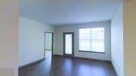 This single-floor space showcases a clean, modern design with a neutral color palette throughout. The main living area is finished with rich, dark hardwood flooring that contrasts attractively against the light gray-white walls, creating a contemporary aesthetic. A large window fitted with white blinds allows abundant natural light to fill the room, while a glass-panel exterior door with matching blinds provides both additional light and direct outdoor access. The smooth white ceilings complete the bright, open feel of the space. An open doorway connects the main area to an adjoining room, suggesting a flexible, multi-room layout well-suited for a variety of uses. The trim and door frames are finished in a dark tone that complements the flooring, lending a cohesive and polished look throughout. The overall ambiance is clean, understated, and move-in ready, reflecting a straightforward modern design approach with practical features. The space benefits from its single-floor layout, making it easily accessible and functional, with a well-balanced interplay of natural light and neutral finishes.