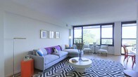This single-floor space offers a thoughtfully designed living area that balances modern aesthetics with comfortable functionality. The living room serves as the centerpiece, anchored by a generous sectional sofa in a soft blue-grey tone, accented with bold purple and blue throw pillows that add a vibrant pop of color. A sleek, round white coffee table sits atop a striking black-and-white geometric area rug, lending a contemporary edge to the space. Framed artwork adorns the clean white walls, complementing the neutral palette throughout. A defining architectural feature is the expansive floor-to-ceiling window wall, fitted with large dark-framed glass panels that flood the interior with natural light and frame lush, tree-lined outdoor views. The seating area near the windows includes elegant armchairs arranged to take full advantage of the scenery, making it an ideal spot for relaxation. Light hardwood flooring runs throughout, enhancing the sense of openness and warmth. The overall ambiance is bright, refined, and inviting, with a cohesive design that blends clean lines, carefully chosen furnishings, and an abundance of natural light into a polished and comfortable living environment.