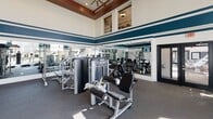 This fitness center on Northlake Centre Pkwy in Charlotte, NC, is a thoughtfully designed, single-floor space that offers a comprehensive workout environment. The room is distinguished by its dramatically high ceilings, which lend an open, airy feel and are accentuated by clerestory windows positioned near the top of the walls, allowing additional natural light to filter in from above. Bold navy and white horizontal stripes run along the walls, giving the space a dynamic, athletic aesthetic.

Large floor-to-ceiling windows along one side of the room flood the cardio area with ample natural light and offer views of the surrounding community. This section houses a row of treadmills, ellipticals, and other cardio machines, all equipped with individual monitors. Full-length mirrors line one wall of the strength training area, visually expanding the space while allowing users to monitor their form across a variety of selectorized weight machines and cable systems.

A dedicated functional training zone features a wall-mounted rack stocked with medicine balls and kettlebells, a heavy punching bag, and a functional trainer cable machine. A wall-mounted television serves as an added amenity. Carpet flooring throughout provides cushioning underfoot, and a ceiling fan contributes to ventilation, completing this well-appointed fitness facility.