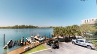 This single-story waterfront property offers an exceptional coastal living experience defined by its prime position along a tranquil marina. The exterior showcases stunning panoramic views of shimmering blue water, with lush tropical palm trees framing the scenery and lending the setting a relaxed, resort-like atmosphere. A private dock area accommodates multiple vessels, making this property an ideal retreat for boating and water sports enthusiasts. The docking facility is well-appointed with sturdy wood-planked piers and secure mooring posts, reflecting careful attention to waterfront infrastructure. On-site parking is conveniently located adjacent to the dock access, providing easy transition between land and water. Across the water, low-rise structures and additional waterfront properties are visible on the opposite shore, reinforcing the sense of a vibrant yet serene coastal community. Clear blue skies overhead and the open water horizon contribute to the bright, airy ambiance that defines this property. Whether arriving by boat or by land, this waterfront space presents an inviting and well-maintained environment that captures the essence of relaxed Florida waterfront living.