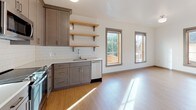 This thoughtfully designed single-floor space combines modern aesthetics with functional living. The kitchen is a standout feature, equipped with gray shaker-style cabinetry accented by brushed gold hardware, stainless steel appliances including a range, microwave, and dishwasher, and a classic white subway tile backsplash. Open natural wood shelving adds warmth and practical storage, while light hardwood flooring runs seamlessly throughout, reflecting an abundance of natural light that pours in through multiple large windows with wood-trimmed frames. The adjacent living area is bright and open, with clean white walls and a generous footprint that offers flexibility for a variety of layout configurations. The bedroom is a calm, sun-lit retreat, featuring the same warm hardwood flooring and a sliding door that contributes to the space's clean, modern feel. The bathroom is finished with a gray vanity topped with a white countertop, a standard toilet, and a towel bar, all set against neutral walls and light flooring. Sleek black hardware on the doors throughout the space provides a subtle contemporary contrast. Overall, the unit presents a cohesive, minimalist design palette that balances practicality with modern style.