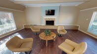 Located at 7000 Red Fox Trail in Shreveport, Louisiana, this single-floor space offers a warm and welcoming layout with thoughtfully designed communal areas. The lounge area serves as a central gathering point, anchored by a tile-surround fireplace with a classic white mantle and a wall-mounted flat-screen television set against a soft blue accent wall. Rich dark hardwood flooring runs throughout, complemented by neutral warm-toned walls and elegant crown molding. Comfortable swivel lounge chairs are arranged around a rustic wooden coffee table, creating an inviting conversational setting. Large windows dressed with horizontal blinds flood the lounge with natural light and offer views of the surrounding greenery. The open dining area features a substantial farmhouse-style trestle table with matching benches, ideal for group gatherings. Adjacent to the dining area, the kitchen is equipped with dark cabinetry, a countertop with bar seating, and modern appliances. A separate windowed room with vaulted beam ceilings and additional large windows provides further flexibility as a multipurpose space. Throughout the space, consistent hardwood flooring, recessed lighting, and crown molding details contribute to a cohesive, polished aesthetic.