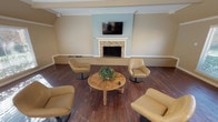 Located at 7000 Red Fox Trail in Shreveport, Louisiana, this single-floor space offers a warm and welcoming layout with thoughtfully designed communal areas. The lounge area serves as a central gathering point, anchored by a tile-surround fireplace with a classic white mantle and a wall-mounted flat-screen television set against a soft blue accent wall. Rich dark hardwood flooring runs throughout, complemented by neutral warm-toned walls and elegant crown molding. Comfortable swivel lounge chairs are arranged around a rustic wooden coffee table, creating an inviting conversational setting. Large windows dressed with horizontal blinds flood the lounge with natural light and offer views of the surrounding greenery. The open dining area features a substantial farmhouse-style trestle table with matching benches, ideal for group gatherings. Adjacent to the dining area, the kitchen is equipped with dark cabinetry, a countertop with bar seating, and modern appliances. A separate windowed room with vaulted beam ceilings and additional large windows provides further flexibility as a multipurpose space. Throughout the space, consistent hardwood flooring, recessed lighting, and crown molding details contribute to a cohesive, polished aesthetic.