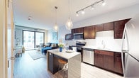 This thoughtfully designed single-floor residence showcases an open-concept layout that seamlessly connects the kitchen, dining, and living areas into one cohesive, flowing space. The kitchen is a standout feature, appointed with rich dark wood cabinetry, granite countertops, and a full suite of stainless steel appliances, including a gas range, microwave, and dishwasher. A crisp white subway tile backsplash adds a clean, modern contrast to the warm cabinetry tones. A central island with a granite surface and bar seating serves as both a functional workspace and an informal dining spot, illuminated overhead by a pair of elegant glass pendant lights. Track lighting along the kitchen ceiling provides additional task illumination. Wide-plank hardwood-style flooring flows throughout the space, unifying the open floor plan. The living area is bright and welcoming, anchored by large floor-to-ceiling windows and a sliding glass door that bathe the interior in natural light while offering views of the surrounding urban environment. The overall ambiance is contemporary and well-appointed, blending warm wood tones, cool gray accents, and modern fixtures to create a stylish and functional living environment.