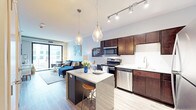 This thoughtfully designed single-floor residence showcases an open-concept layout that seamlessly connects the kitchen, dining, and living areas into one cohesive, flowing space. The kitchen is a standout feature, appointed with rich dark wood cabinetry, granite countertops, and a full suite of stainless steel appliances, including a gas range, microwave, and dishwasher. A crisp white subway tile backsplash adds a clean, modern contrast to the warm cabinetry tones. A central island with a granite surface and bar seating serves as both a functional workspace and an informal dining spot, illuminated overhead by a pair of elegant glass pendant lights. Track lighting along the kitchen ceiling provides additional task illumination. Wide-plank hardwood-style flooring flows throughout the space, unifying the open floor plan. The living area is bright and welcoming, anchored by large floor-to-ceiling windows and a sliding glass door that bathe the interior in natural light while offering views of the surrounding urban environment. The overall ambiance is contemporary and well-appointed, blending warm wood tones, cool gray accents, and modern fixtures to create a stylish and functional living environment.