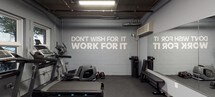 Located at 619-633 Adams St in Hoboken, New Jersey, this fitness center is a fully outfitted single-floor gym designed to support a wide range of workout routines. The space features a clean, industrial aesthetic with painted concrete block walls in a cool grey palette, exposed ceiling infrastructure, and durable rubber flooring throughout — all contributing to a focused, no-frills training environment. A bold motivational phrase, "Don't Wish For It, Work For It," is prominently displayed on the wall, setting an energizing tone for the space. A large floor-to-ceiling mirror along one wall visually expands the room while allowing users to monitor their form. Cardio equipment includes multiple treadmills and an elliptical machine, as well as a Peloton stationary bike with an interactive screen. Strength training is well represented with a multi-function cable and weight machine, a flat workout bench, dumbbells, and resistance bands mounted on the wall. Stability balls are neatly stored on a wall-mounted rack, and a wall-mounted television provides additional entertainment options. Natural light filters in through several windows, and a wall-mounted HVAC unit ensures climate control. The space is accessible via a dedicated entry door, with an emergency exit clearly marked.