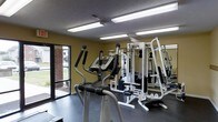 This single-floor fitness center is a dedicated workout space designed to accommodate a range of exercise needs. The room features a functional layout with a variety of equipment, including an elliptical trainer and a multi-station cable weight machine, providing users with both cardiovascular and strength training options. The space is illuminated by bright overhead fluorescent lighting, ensuring excellent visibility throughout the room. A large window along one wall allows natural light to filter in, offering a pleasant view of the surrounding brick exterior and greenery outside. The walls are painted in a warm yellow-green tone, contributing to an energizing atmosphere, while the dark rubberized flooring provides a durable, slip-resistant surface well-suited for an active environment. The textured ceiling adds a utilitarian character consistent with the space's purpose. Overall, this fitness center delivers a practical and well-equipped environment for residents or users looking to maintain an active lifestyle, with its thoughtful combination of natural and artificial lighting creating a welcoming and functional workout space.