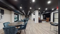 This single-floor communal amenity space is designed with a contemporary aesthetic that balances striking contrast and refined comfort throughout its open layout. A dramatic matte black ceiling anchors the entire space, complemented by recessed lighting that casts a warm, focused glow across the light wood-look tile flooring below. Crisp white walls and structural columns provide visual balance, preventing the darker palette from overwhelming the interior.

The space is organized into several distinct yet interconnected zones. A dining area features round tables paired with plush teal velvet chairs, offering a casual yet elevated setting for gatherings. Adjacent to this, a lounge area is outfitted with a large sectional sofa in deep charcoal, leather accent chairs, and a substantial wooden coffee table, all arranged in front of a large wall-mounted media screen—ideal for communal viewing.

A dedicated kitchen wet bar runs along one wall, fitted with dark cabinetry, patterned tile backsplash, pendant lighting, and stainless steel appliances, providing full functionality for entertaining. Black-framed glass double doors lead directly to an outdoor patio, introducing natural light and seamless indoor-outdoor connectivity. The overall ambiance is modern, sophisticated, and purposefully designed for social engagement.