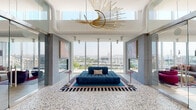 This elegantly designed amenity floor offers a dynamic, multi-zone social experience within a contemporary high-rise setting. The space is anchored by a dramatic central lounge featuring polished terrazzo flooring, floor-to-ceiling curtain-wall glazing, and a bold sculptural gold chandelier overhead. A deep-tufted royal blue sofa sits beneath large-scale colorful artwork and a two-sided fireplace, framing panoramic views of the city skyline — including a prominent domed civic landmark visible in the distance.

Adjacent seating lounges continue the floor-to-ceiling window theme, furnished with curated collections of plush velvet and sculptural chairs in rich jewel tones — deep purples, magentas, and warm neutrals — set over abstract area rugs. Each zone is thoughtfully arranged to maximize the sweeping urban views while maintaining an intimate, lounge-like atmosphere.

A dedicated games room features light hardwood flooring, a full-size billiards table, built-in banquette seating with a bar-height counter, pendant lighting, and a wall-mounted television. A well-appointed catering kitchen with white quartz countertops, dark cabinetry, a textured geometric tile feature wall, and a large mounted display screen supports the entertaining areas. Dual stainless steel elevators with terrazzo flooring complete the arrival experience.