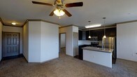 This single-floor space offers an open-concept layout that seamlessly connects the living area and kitchen, creating a sense of spaciousness and flow. The living area is finished with wall-to-wall carpet and features a ceiling fan with a light fixture, adding both comfort and functionality overhead. Crown molding details along the upper walls lend a subtle touch of architectural refinement throughout the space.

The kitchen is a standout feature, designed with rich, dark cabinetry that provides striking contrast against the light-toned walls. A kitchen island with a granite-style countertop and an integrated sink serves as both a functional workspace and a natural gathering point. Stainless steel appliances — including a refrigerator, range, and over-the-range microwave — are neatly integrated into the cabinetry. Pendant lighting hangs above the island, complementing the overhead fluorescent fixture that ensures the kitchen is well-lit.

A hallway visible from the living area leads further into the space, suggesting additional rooms beyond the main open area. The transition from carpeted living space to hard flooring in the kitchen adds a practical and visually defined separation between the two areas. Overall, the space presents a clean, modern, and functional interior with a cohesive dark-and-neutral color palette.
