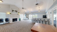 This thoughtfully designed single-floor community amenity space offers a generous layout encompassing a lounge, fitness center, meeting room, and a kitchen bar area. The lounge is a welcoming centerpiece, featuring vaulted ceilings, patterned carpeting, and a striking stone-surround fireplace flanked by decorative artwork and warm accent lighting. Pendant lights hang from the soaring ceiling, adding a stylish touch, while large windows flood the space with natural light. A comfortable seating area completes this relaxing gathering spot. Adjacent to the lounge is a granite-topped kitchen bar with ample seating for bar-height guests, stainless-accented metal stools, and direct access to an outdoor balcony through glass-paned French doors, offering views of surrounding greenery. A dedicated meeting area provides flexible table and chair arrangements, enhanced by natural light from large windows and tasteful décor including a classic wall clock and wooden credenza. The fitness center is well-appointed with a variety of cardio equipment including treadmills, ellipticals, a stationary bike, and a rowing machine, all set on durable dark flooring with multiple windows providing natural light and ventilation. Together, these spaces create a functional and inviting community hub suited for socializing, meetings, and active living.