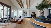 This single-story amenity and clubhouse space is defined by its striking architectural design and carefully curated interior. The entry foyer sets an immediate tone with bold black-and-white checkered marble-style tile flooring, black-framed glass doors, and wall sconces that frame views of the palm-tree-lined exterior. A round glass-top accent table anchors the entry, creating a refined first impression.

The space transitions into a grand main lounge area distinguished by soaring vaulted ceilings with exposed wood beam detailing and warm herringbone-patterned hardwood flooring. A dramatic circular reception or display platform, crafted in wood and dark cabinetry, anchors the room's center and hosts a live olive tree, adding a natural focal point. Comfortable seating arrangements with neutral upholstered sofas, leather ottomans, and layered rugs invite gathering, while brass pendant lighting and wall-mounted art contribute to a sophisticated coastal ambiance.

A floor-to-ceiling arched window wall offers panoramic views of the outdoor pool area and palm trees, flooding the lounge with natural light. An adjacent game and social room features a pool table, bar seating, a chandelier, and large windows, providing a more casual entertainment setting throughout.
