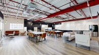 This single-story commercial space showcases a striking industrial-modern aesthetic that blends raw architectural elements with refined finishes. The open floor plan is anchored by soaring exposed ceilings featuring bold red steel trusses, visible ductwork, and a combination of rectangular LED light fixtures and Edison-style pendant bulbs that create a warm, layered lighting scheme throughout.

Light hardwood flooring runs seamlessly across the entire space, unifying the multiple functional zones within. The main area is configured with large white marble-topped communal tables and cognac leather chairs, providing ample seating for collaborative work or group gatherings. A lounge area offers plush white sofas and casual seating arrangements for more relaxed interactions.

The kitchen area is a standout feature, equipped with a full-length run of gray shaker-style cabinetry, a dramatic floor-to-ceiling white marble slab backsplash, a large waterfall-edge island with bar seating, stainless steel appliances including a refrigerator and beverage cooler, and a deep undermount sink. An exposed brick wall adds warmth and texture adjacent to the kitchen. Dark vertical shiplap accent walls and frosted glass-paneled enclosures provide visual contrast, while multiple windows allow natural light to filter throughout the space.