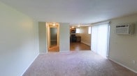 This single-floor unit offers a straightforward, functional layout with a clear distinction between the living and kitchen areas. The living room is finished with neutral-toned carpet and light green walls, creating a calm, understated atmosphere. A large window fitted with vertical blinds along one wall allows natural light to filter into the space, brightening the interior throughout the day. A baseboard heating unit runs along the wall, providing an efficient source of warmth.

The living area opens directly into the kitchen, which transitions to hardwood-style flooring. The kitchen is equipped with black appliances, including a range and microwave, set against warm wood cabinetry. A window above the counter area brings additional natural light into the cooking space. The kitchen also features its own baseboard heater, ensuring consistent comfort throughout the unit.

A hallway visible from the main living area leads to additional rooms, with a frosted-glass door providing privacy. Flush-mount ceiling lights are installed throughout, offering consistent, even illumination across all areas of the space. The overall design is clean and practical, well-suited for everyday living.