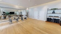 Located at 1 Main St in Miramar, FL, this contemporary fitness studio is a thoughtfully designed, single-floor space that delivers a well-rounded workout environment. The studio features warm-toned hardwood flooring throughout, crisp white walls, and recessed lighting that creates a clean, energizing atmosphere.

The space is divided into distinct functional zones. A dedicated cycling area houses a row of stationary spin bikes positioned in front of a large, floor-to-ceiling mirror fitted with a ballet barre, making it equally suited for barre-style workouts. Opposite, a functional training zone features a multi-station exercise rack stocked with medicine balls and resistance bands, offering versatile strength and conditioning options.

A serene meditation and yoga corner provides a calming contrast, outfitted with open shelving displaying candles, greenery, and decorative figurines, alongside neatly stored yoga mats and cushions. Large framed mirrors in this zone visually expand the space and support movement practice.

Expansive windows with horizontal louvers line the upper walls, flooding the studio with natural light and offering views to an outdoor area. Sheer roller shades allow for light control while maintaining an airy feel. The studio's modern design, multi-use layout, and abundant natural light make it a versatile and inviting fitness destination.