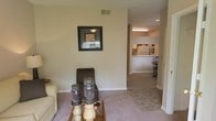 This single-story residence presents a straightforward, functional layout with an open flow between the main living area and the kitchen. The living room is carpeted and features neutral-toned walls, creating a calm and understated atmosphere. A framed piece of wall art and a glass-top coffee table with decorative candle accents add a touch of personality to the space. Recessed ceiling vents and standard ceiling height contribute to a practical, well-maintained interior.

A wide open passageway connects the living room to the kitchen, which is visible from the main seating area. The kitchen features a raised breakfast bar that serves as a natural divider, along with overhead fluorescent lighting that brightens the workspace. White appliances, including a refrigerator, are visible within the kitchen, and upper cabinetry lines the walls, providing ample storage. A small dining area is partially visible adjacent to the kitchen, suggesting a convenient eat-in configuration. Overall, the space offers a cohesive, comfortable layout suited for everyday living.