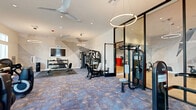 Located at 29986 Berea Road in Menifee, CA, this modern fitness center offers a thoughtfully designed layout across a single floor, divided into two distinct functional zones. The main workout area features patterned carpet tile flooring and is outfitted with a comprehensive selection of strength and cardio equipment, including treadmills, elliptical machines, multi-station cable machines, adjustable benches, and a full dumbbell rack. Suspended circular ring pendant lights and ceiling fans add both style and comfort to the space. Large windows line the perimeter, flooding the area with natural light and providing views of the surrounding community. A bold geometric wall mural adds an artistic focal point to the room, enhancing the contemporary aesthetic. Separated by sleek floor-to-ceiling black-framed glass partition walls, a dedicated studio room offers a change of environment for more varied workouts. This secondary space features warm wood-look flooring, full-length mirrored walls, a mounted flat-screen TV, stationary bikes, rowing equipment, and red exercise mats, making it ideal for cycling, stretching, or guided fitness sessions. A ceiling fan and recessed lighting keep the studio bright and well-ventilated. Together, the two zones create a versatile, modern fitness facility suited for a wide range of workout routines.
