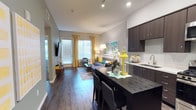 This contemporary single-floor residence is designed with a clean, modern aesthetic and a cohesive color palette of gray, teal, and yellow accents that flows throughout the space. The open-concept layout connects the kitchen and living area seamlessly, creating a bright and functional environment. The kitchen is outfitted with dark espresso cabinetry, white quartz countertops, a subway tile backsplash, and a full suite of stainless steel appliances including a gas range, microwave, dishwasher, and French door refrigerator. A dark-topped kitchen island with bar seating provides additional prep space and casual dining. The living area benefits from large windows dressed with golden yellow curtains that invite natural light, and features wood-look flooring that adds warmth to the space. The bedroom is carpeted for comfort and showcases a sage green accent wall, large windows with patterned drapes, and ample room for a full furniture arrangement. The bathroom continues the modern theme with dark wood vanity cabinetry, a white countertop, wood-look flooring, and a bright yellow accent wall that adds a vibrant focal point. Track lighting and recessed fixtures are present throughout, ensuring well-lit interiors across all areas of the residence.